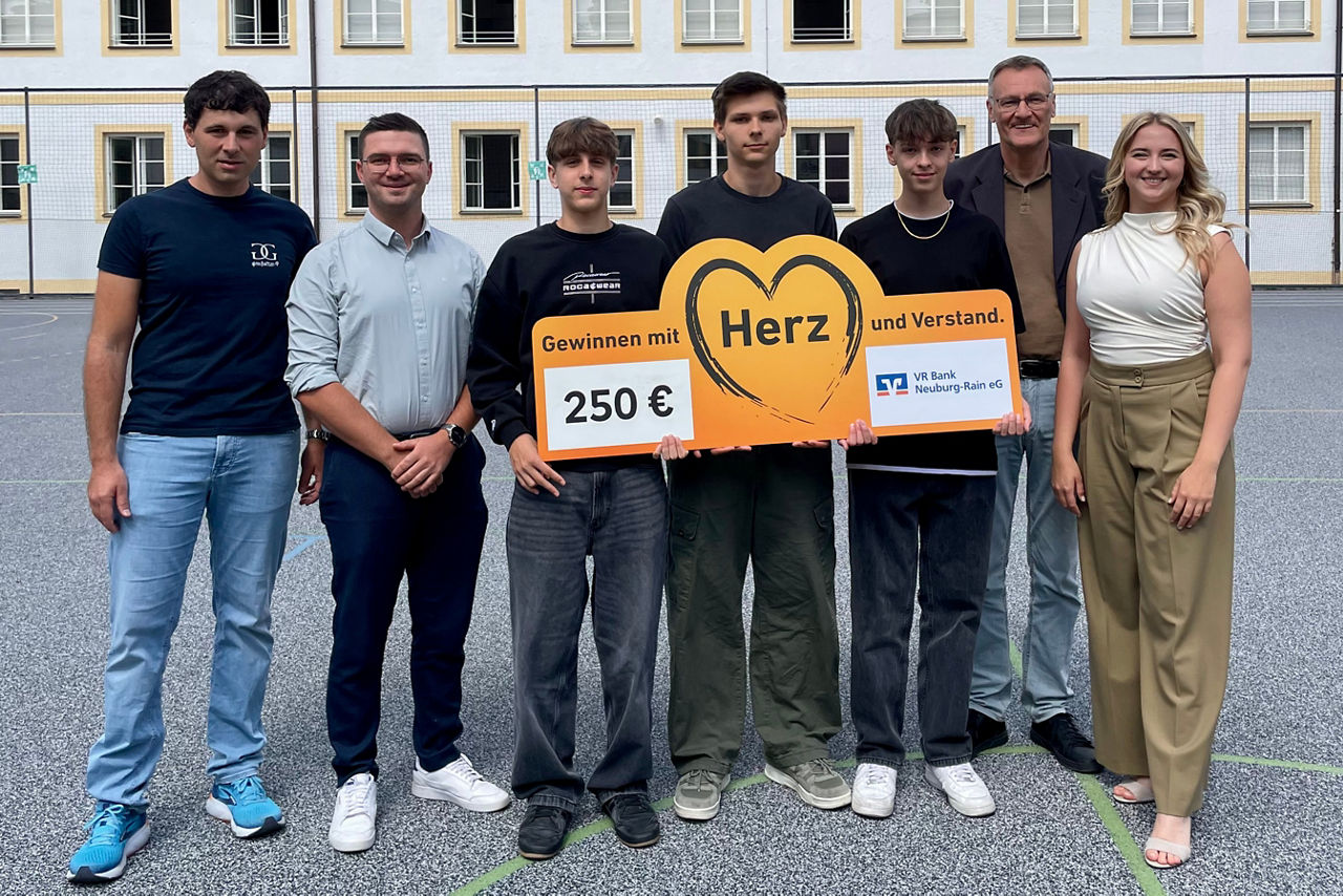 Eine Gruppe von Schülern und Erwachsenen vor einem Schulgebäude halten einen Spendenscheck in den Händen.