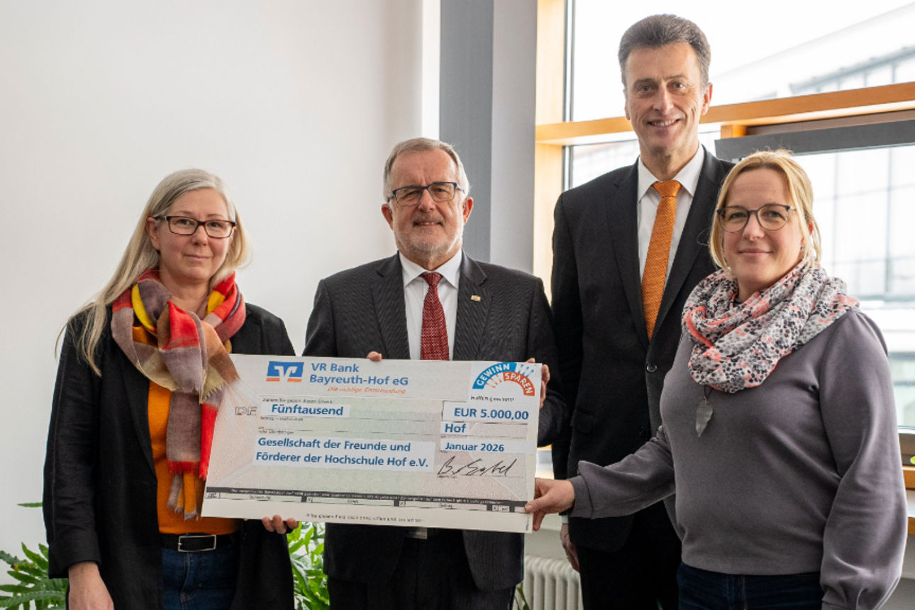 Vier Personen überreichen gemeinsam einen großen symbolischen Spendenscheck in einem Büroraum.