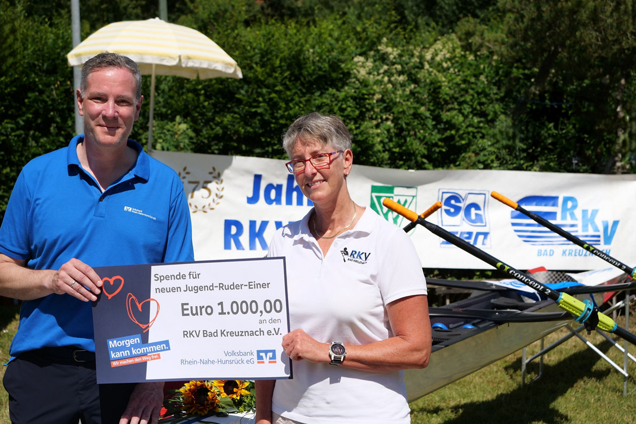 Zwei Personen stehen neben auf einer Wiese und halten in der Mitte gemeinsam einen Spendenscheck.