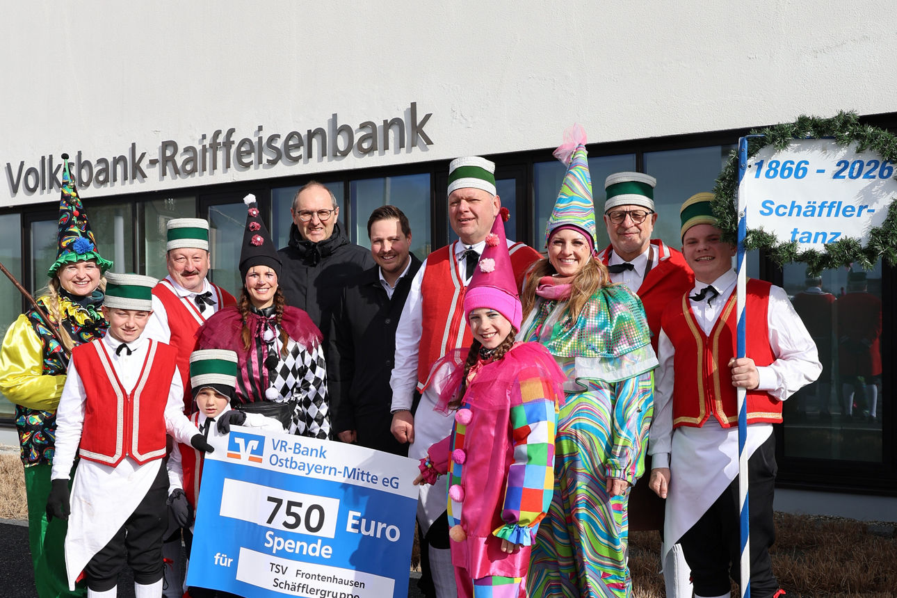 Gruppe von elf Personen in bunten Karnevalskostümen und Vereinskleidung vor einem Bankgebäude