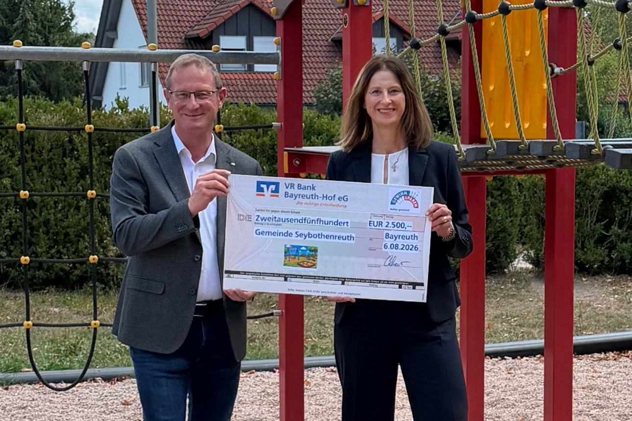Zwei Personen stehen auf einem Spielplatz vor bunten Klettergeräten und präsentieren einen symbolischen Spendenscheck.
