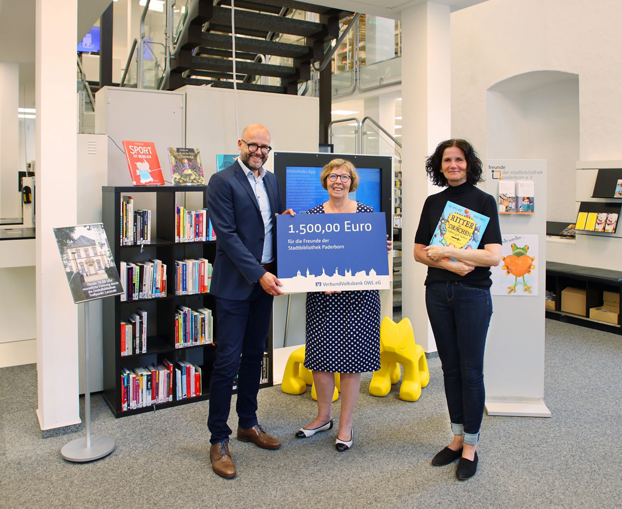 Drei Personen bei der symbolischen Übergabe einer Spende über 1.500 Euro in der Stadtbibliothek Paderborn.