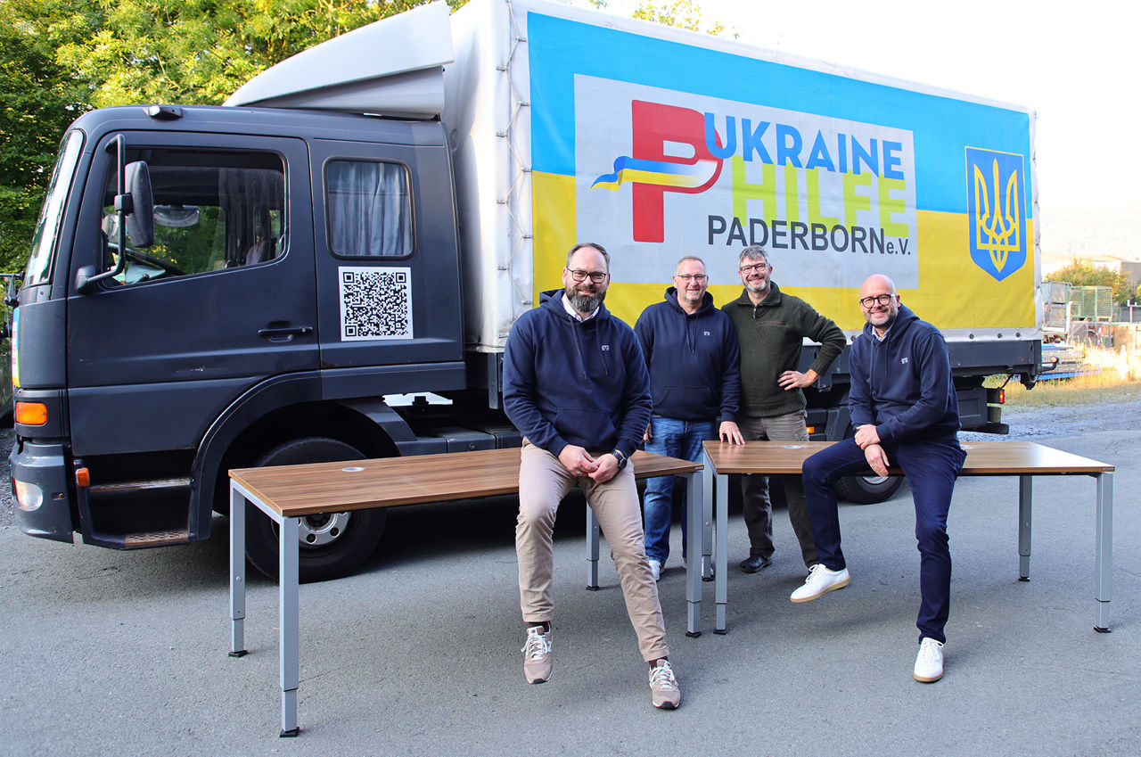 Karsten Stasch von der Ukraine Hilfe Paderborn steht mit Mitarbeitern der VerbundVolksbank OWL vor den gespendeten Schreibtischen und einem LKW.