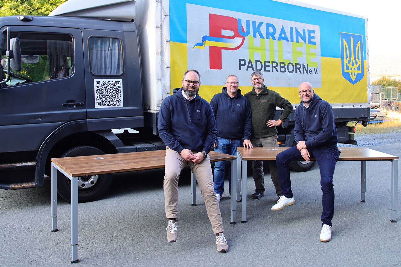 Karsten Stasch von der Ukraine Hilfe Paderborn steht mit Mitarbeitern der VerbundVolksbank OWL vor den gespendeten Schreibtischen und einem LKW.