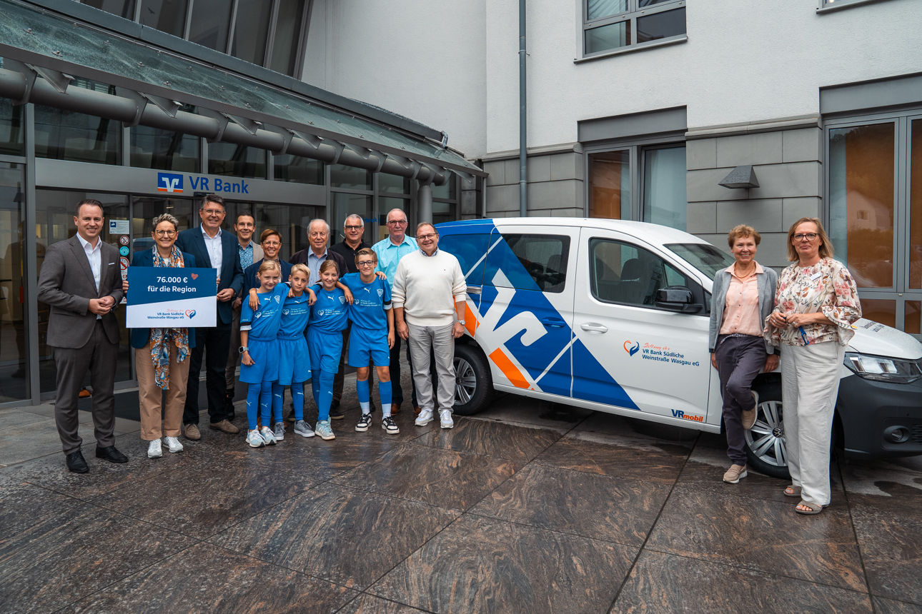 Gruppe vor VR Bank, hält Schild mit ‚7.000 € für die Region‘, daneben Auto mit VR Bank-Logo.