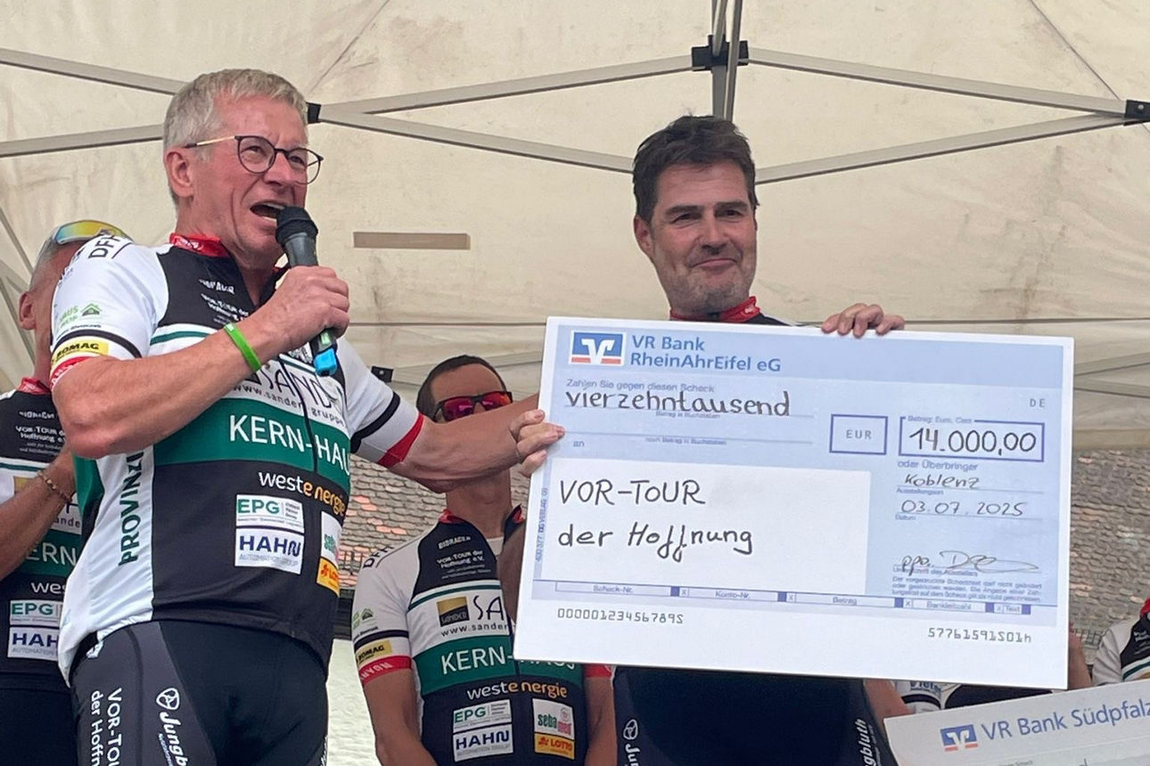 Zwei männliche Personen aus einem Radfahrer-Team halten einen großen symbolischen Spendenscheck in den Händen.