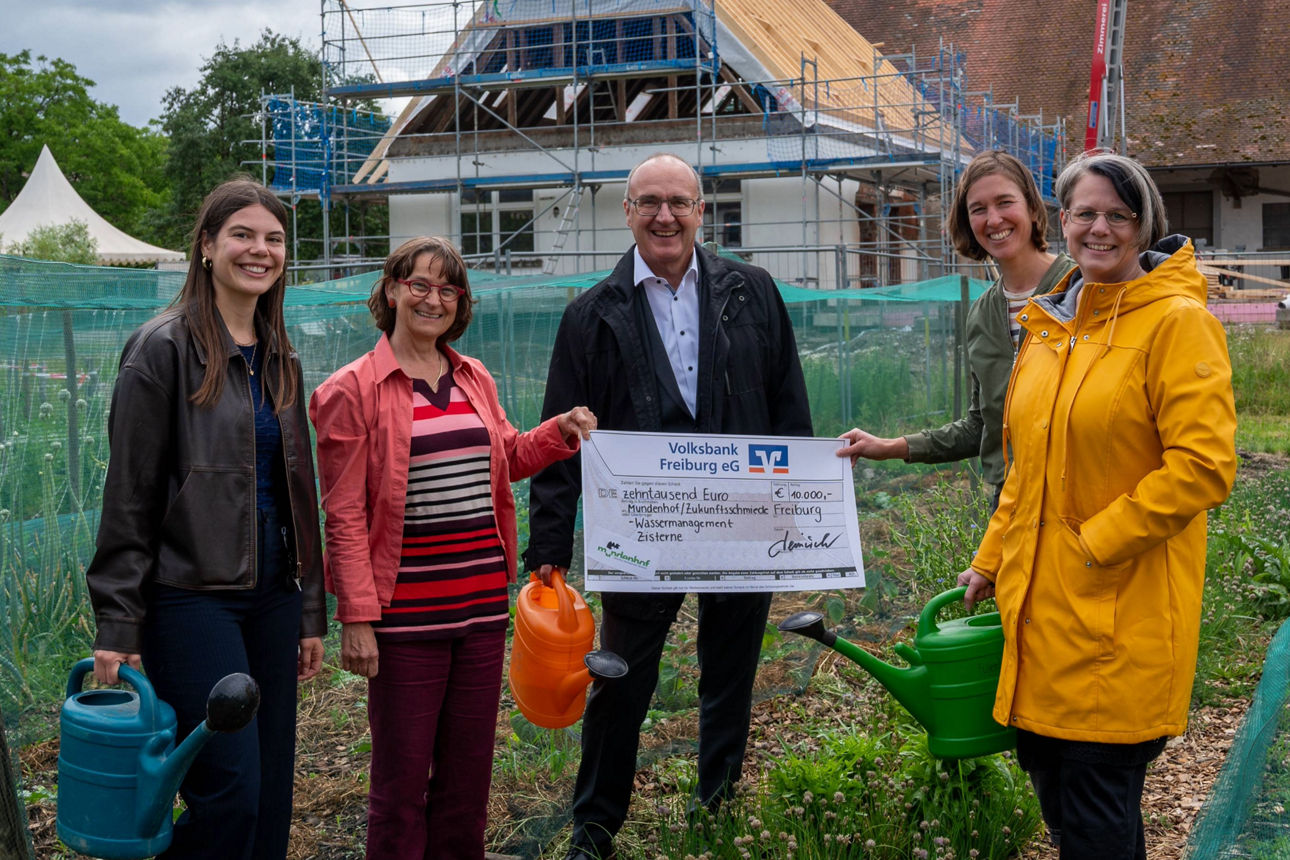 Fünf Personen halten einen Spendenscheck über 10.000 Euro auf einem Gelände mit Neubau und Gießkannen.