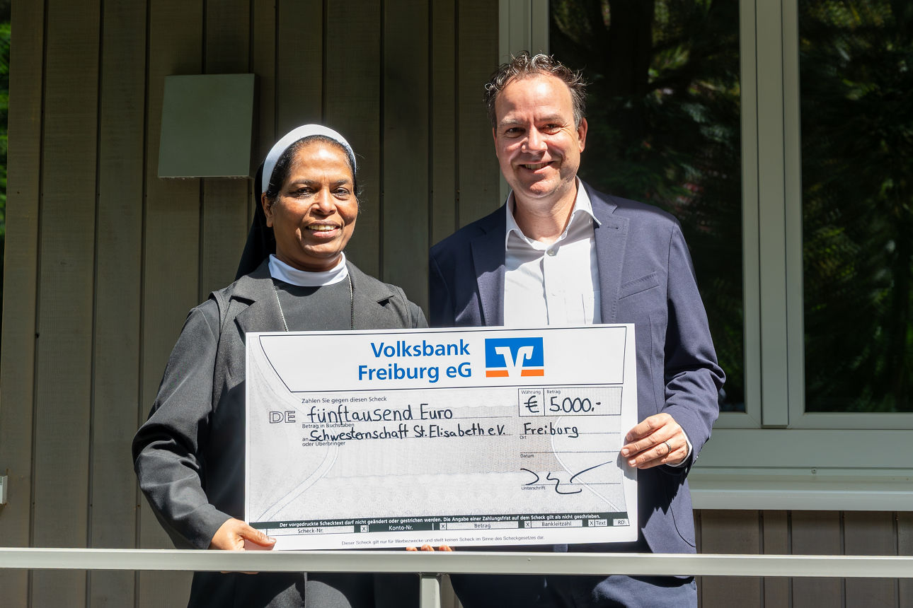 Zwei Personen halten gemeinsam einen großen Spendenscheck über 5.000 Euro vor einem Gebäude im Freien.