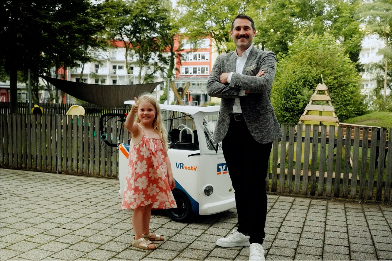 Bankmitarbeiter steht mit Kind vor VR Mobil und lacht in die Kamera