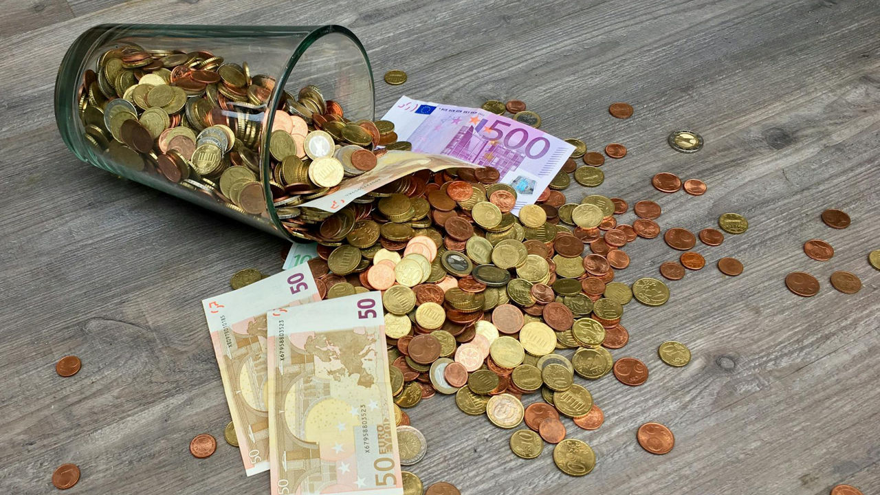Ein Glasgefäß, das mit Münzen überquillt und neben Euro-Banknoten auf eine hölzerne Oberfläche fällt.