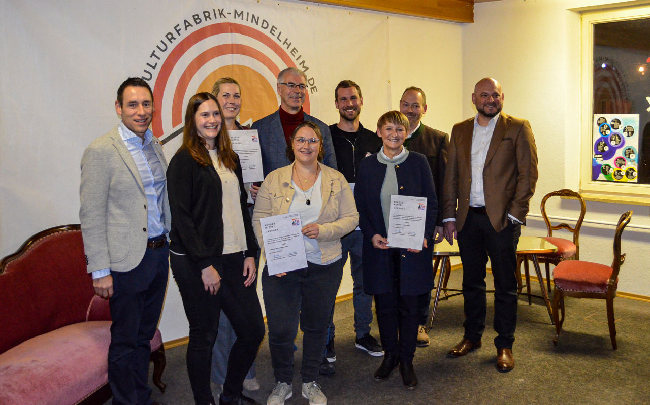 9 Personen stehen in förmlicher Kleidung nebeneinander und lächeln in die Kamera.