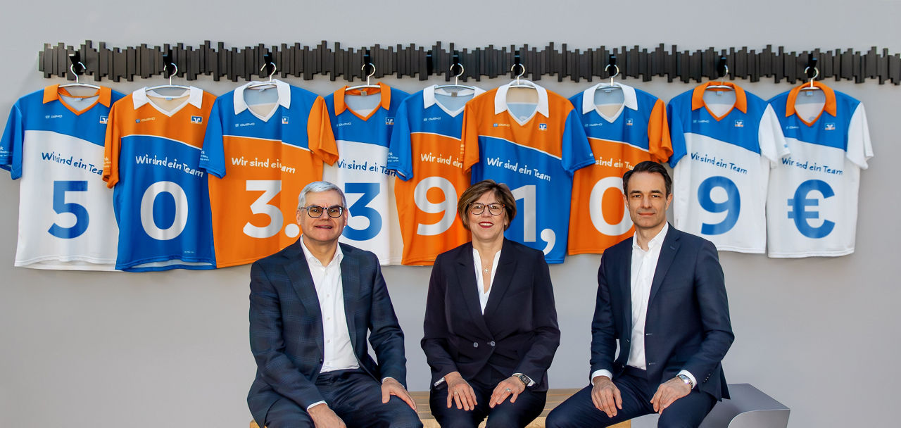 Das Bild zeigt den Vorstand der VR Bank Main-Kinzig-Büdingen vor einer grauen Wand an der neun Trikots in den Farben Blau, Weiß und Orange. Auf jedem Trikot ist eine Zahl abgebildet zusammen ergeben sie 503.391,09€