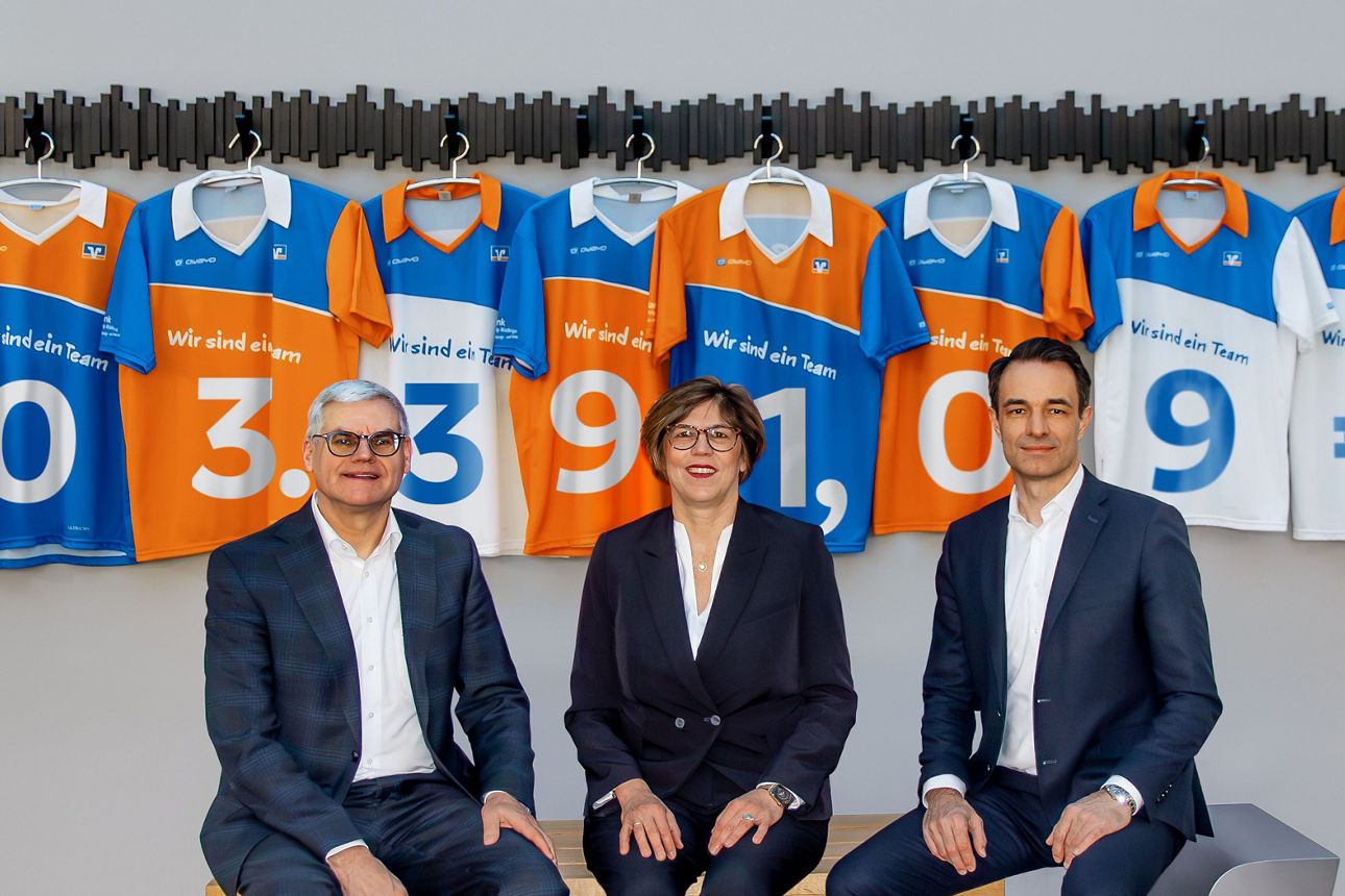 Das Bild zeigt den Vorstand der VR Bank Main-Kinzig-Büdingen vor einer grauen Wand an der neun Trikots in den Farben Blau, Weiß und Orange. Auf jedem Trikot ist eine Zahl abgebildet zusammen ergeben sie 503.391,09€