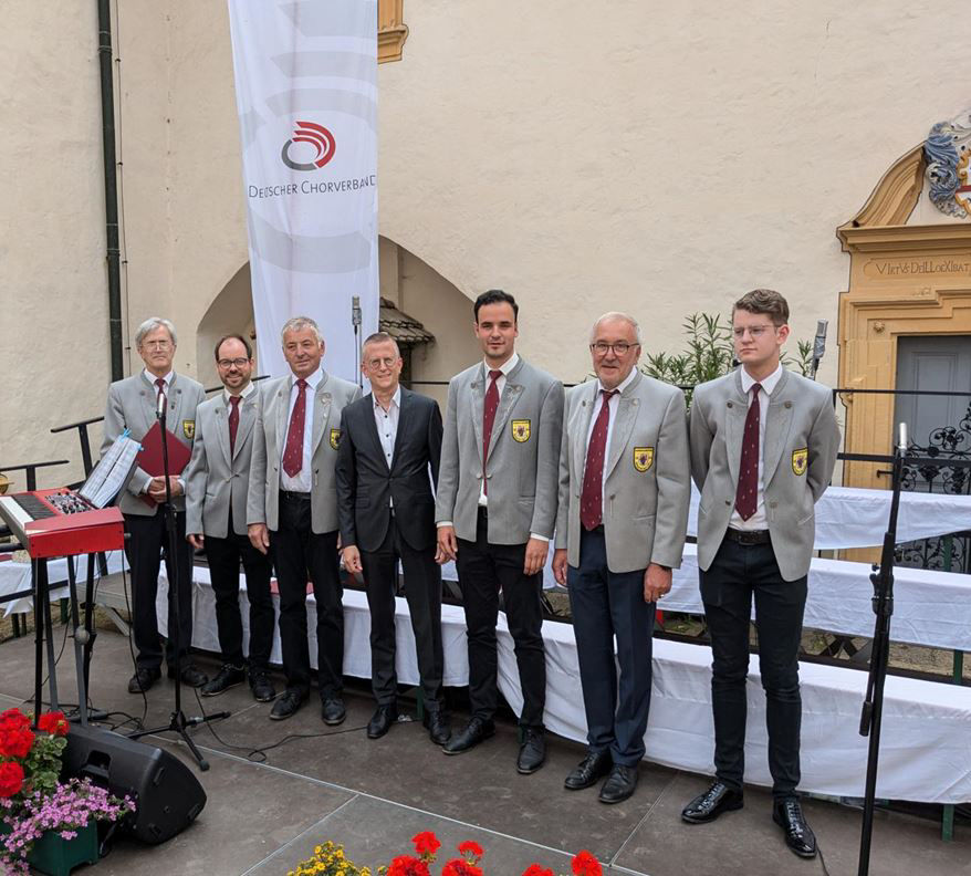 Ein Chor, gekleidet in koordinierten hellgrauen Jacken und Krawatten, steht bereit für einen Auftritt.