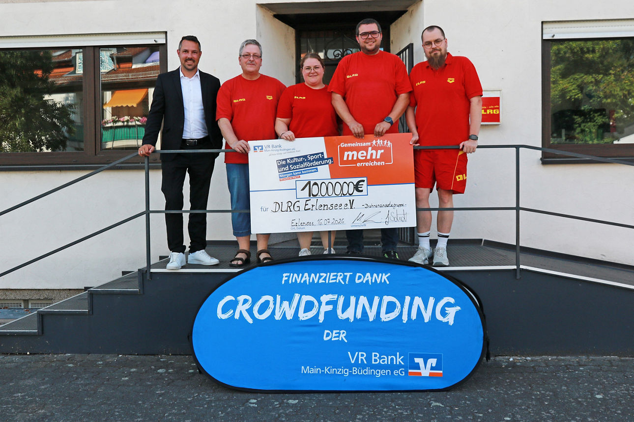 Fünf Personen stehen vor dem Gebäude der DLRG Erlensee und halten einen symbolischen Spendenscheck über 10.000 Euro aus einem Crowdfunding-Projekt der VR Bank Main-Kinzig-Büdingen für die Anschaffung einer Einsatzdrohne.