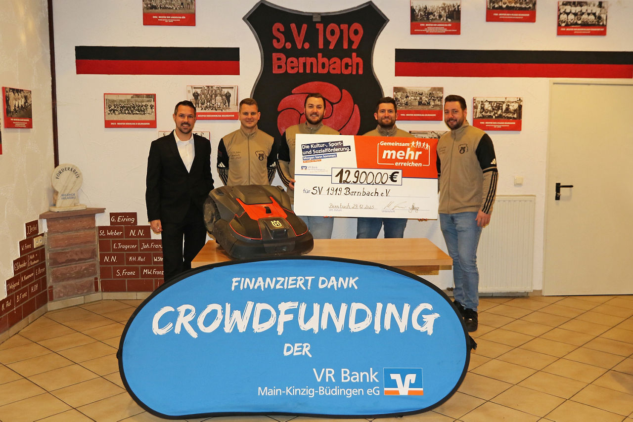 Fünf Männer präsentieren Scheck über 12.900 € und Mähroboter vor Vereinswappen des SV 1919 Bernbach, finanziert durch Crowdfunding der VR Bank.