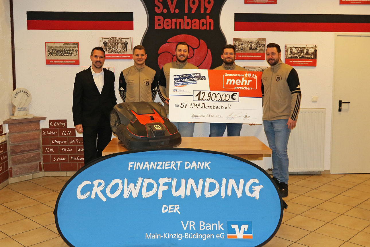 Fünf Männer präsentieren Scheck über 12.900 € und Mähroboter vor Vereinswappen des SV 1919 Bernbach, finanziert durch Crowdfunding der VR Bank.