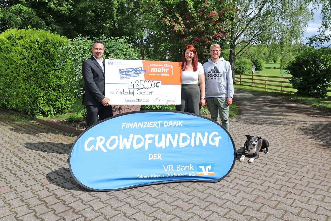 Drei Personen präsentieren einen Scheck über 4.850 € im Freien. Im Vordergrund steht ein blaues Banner mit der Aufschrift „finanziert dank Crowdfunding der VR Bank Main-Kinzig-Büdingen“
