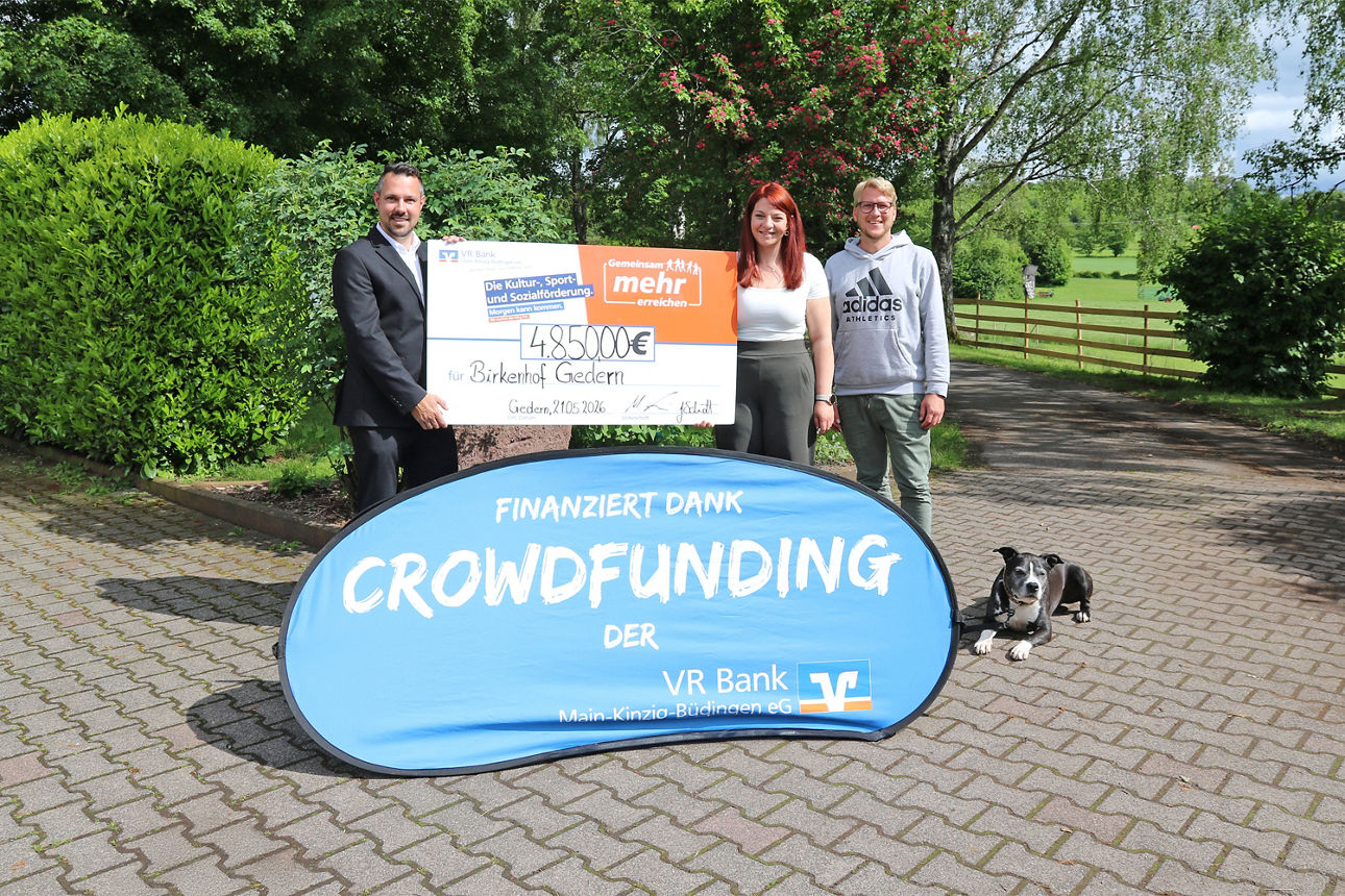 Drei Personen präsentieren einen Scheck über 4.850 € im Freien. Im Vordergrund steht ein blaues Banner mit der Aufschrift „finanziert dank Crowdfunding der VR Bank Main-Kinzig-Büdingen“