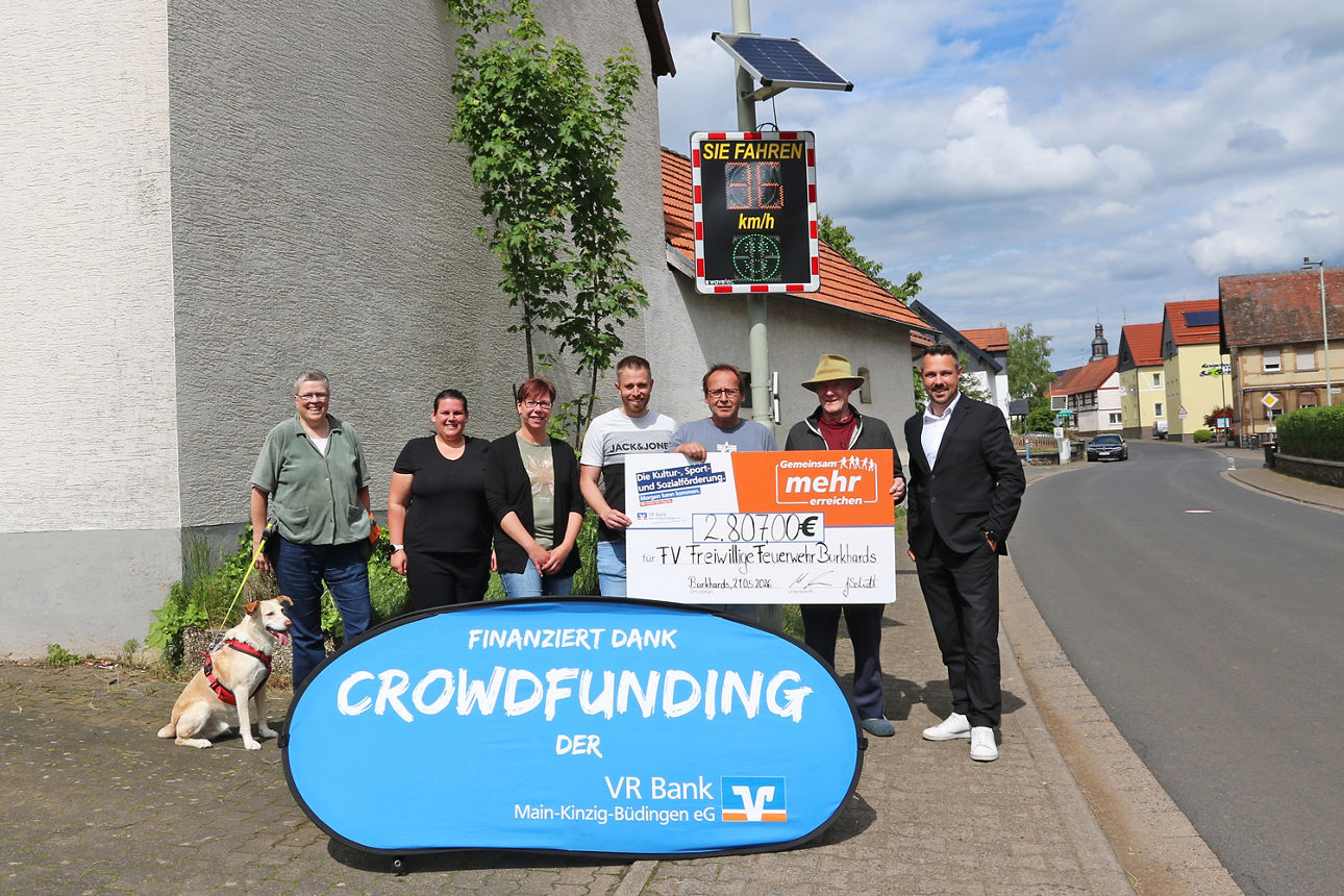 Sieben Personen präsentieren einen Scheck über 2.807 € an einer Straße vor einer Geschwindigkeitsanzeige. Im Vordergrund steht ein blaues Banner mit der Aufschrift „finanziert dank Crowdfunding der VR Bank Main-Kinzig-Büdingen“
