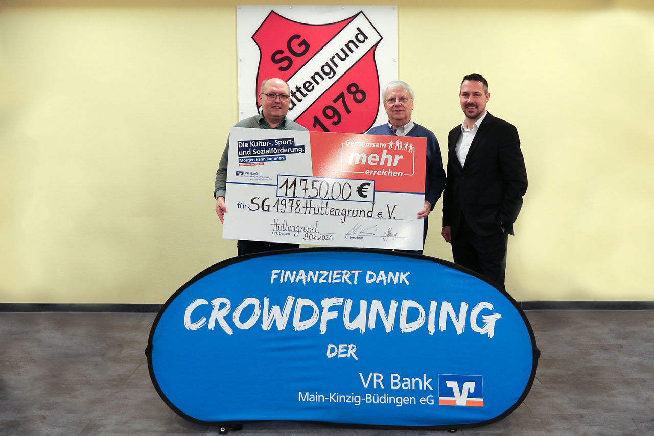 Drei Männer präsentieren einen Scheck über 11.750€ vor dem Wappen der SG Huttengrund 1978. Im Vordergrund ist ein blaues Banner mit der weißen Aufschrift "Finanziert dank Crowdfunding der VR Bank Main-Kinzig-Büdingen eG" zu sehen