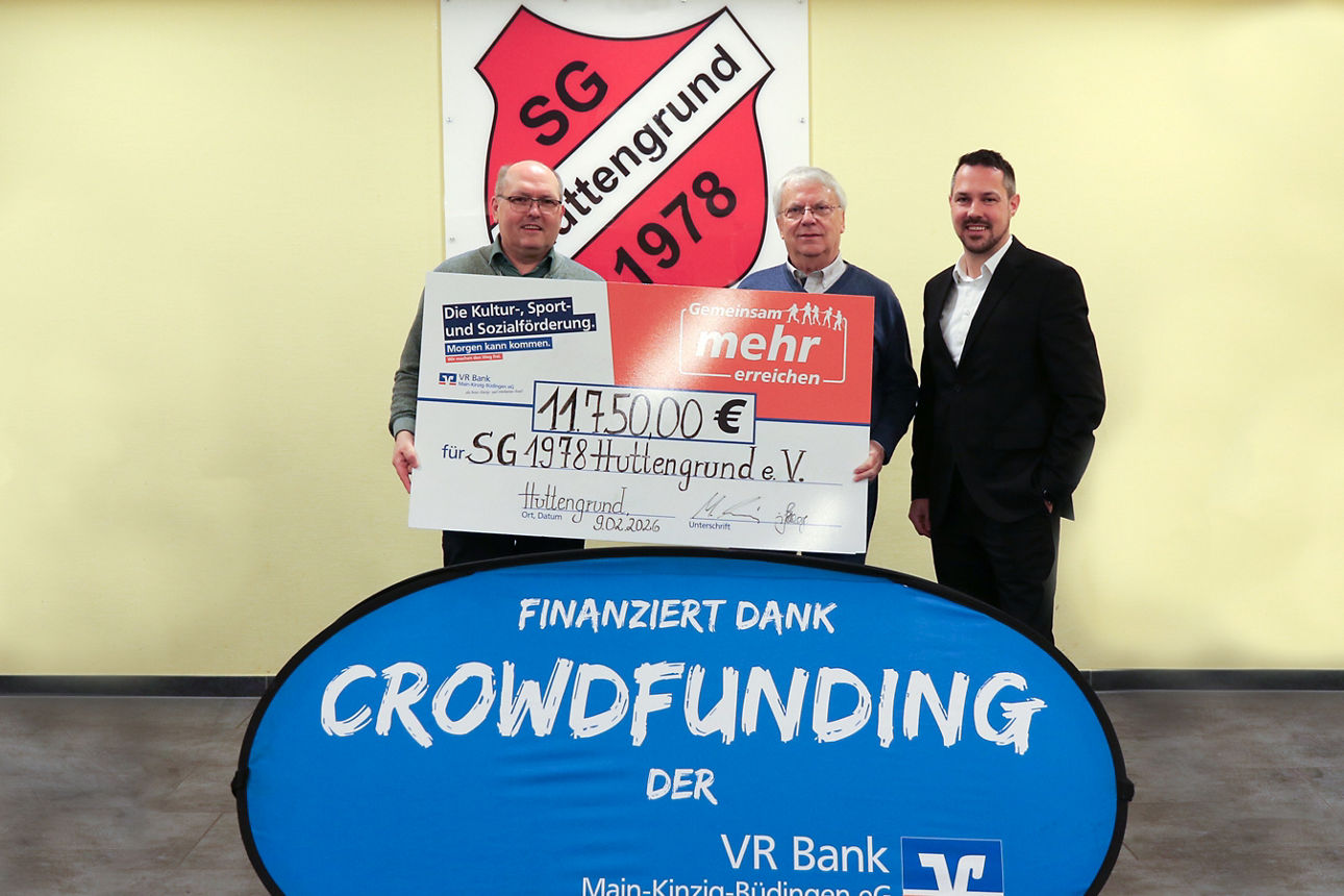 Drei Männer präsentieren einen Scheck über 11.750€ vor dem Wappen der SG Huttengrund 1978. Im Vordergrund ist ein blaues Banner mit der weißen Aufschrift "Finanziert dank Crowdfunding der VR Bank Main-Kinzig-Büdingen eG" zu sehen
