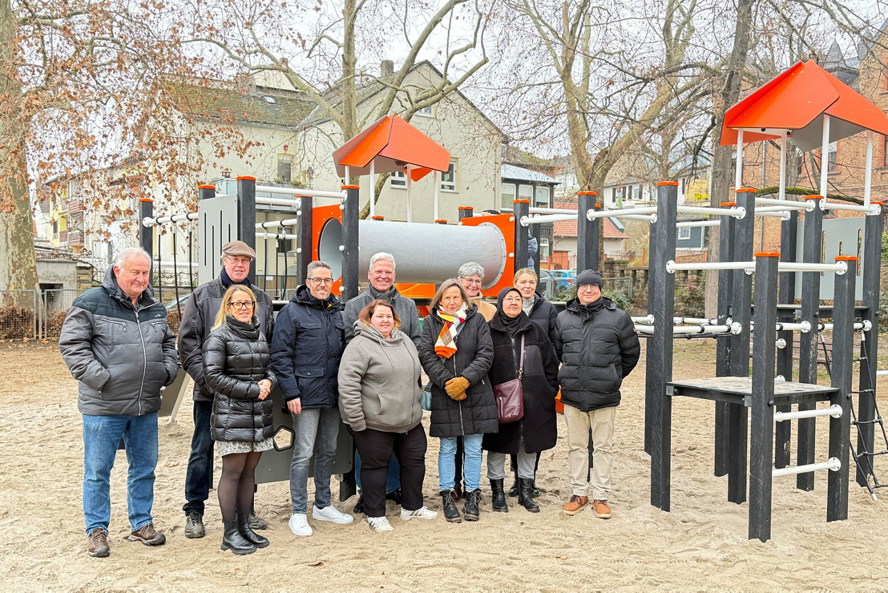 Gruppe von Menschen in Winterkleidung steht vor einem Spielgerät mit Rutschen und Kletterelementen auf einem Sandplatz