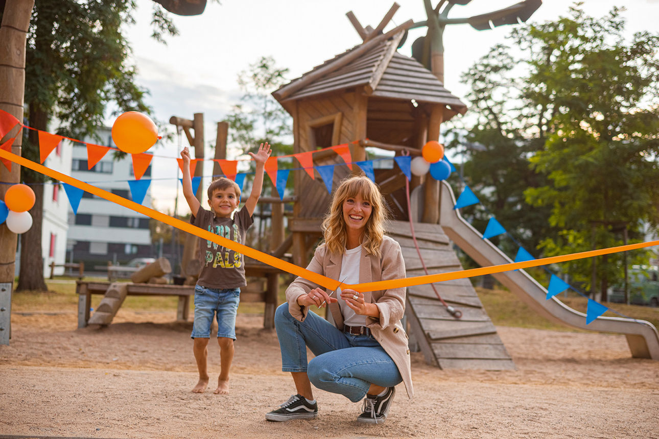 Frau schneidet feierlich ein Band vor einem Spielplatz durch, während ein Junge daneben steht. Bunte Deko im Hintergrund.