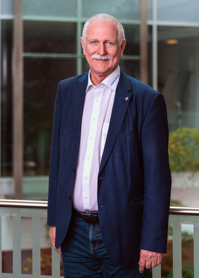 Ein Mann steht im dunklen Anzug vor einer Glaswand und lächelt in die Kamera.