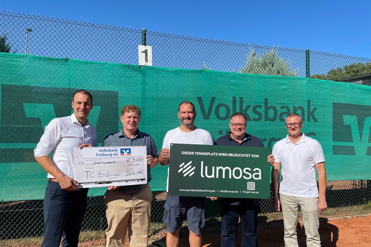 Fünf Personen präsentieren auf einem Tennisplatz einen Spendenscheck und ein Schild zur neuen Platzbeleuchtung.