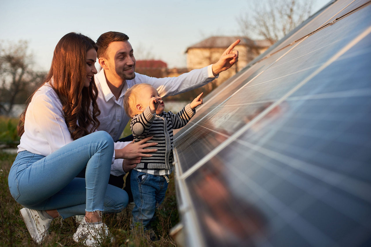 Junge Familie betrachtet gemeinsam eine Solaranlage im Freien bei Sonnenuntergang. Die Eltern knien neben ihrem Kleinkind, das neugierig auf die Solarpanels zeigt.