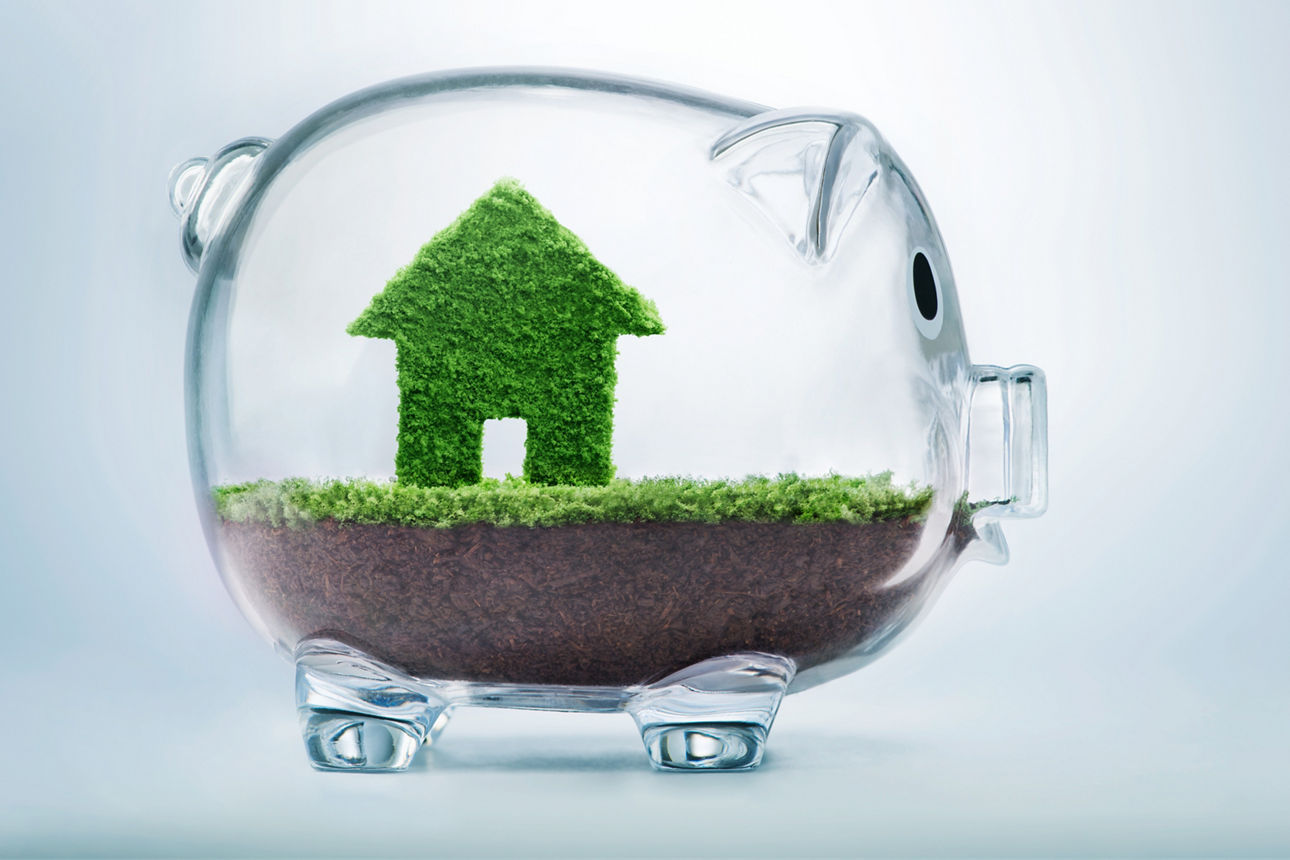 Gläsernes Sparschwein, das mit Erde und Haus aus grünen Bättern gefüllt ist