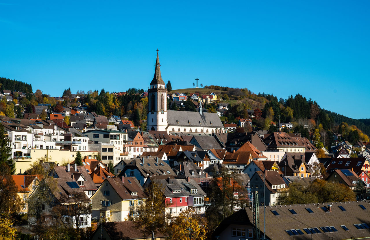 Panoramablick über eine Stadt mit markanter Kirche, bunten Häusern und bewaldeten Hügeln unter klarem Himmel.