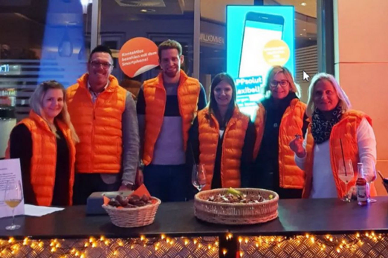 Mehrere Personen in orangefarbenen Westen stehen gemeinsam an einem Aktionsstand vor einem beleuchteten Eingangsbereich.