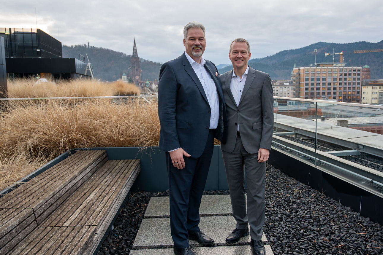 Hr. Brinkmann und Hr. Stahl stehen auf unserer Dachterrasse mit Glasgeländer, im Hintergrund Freiburger Münster.