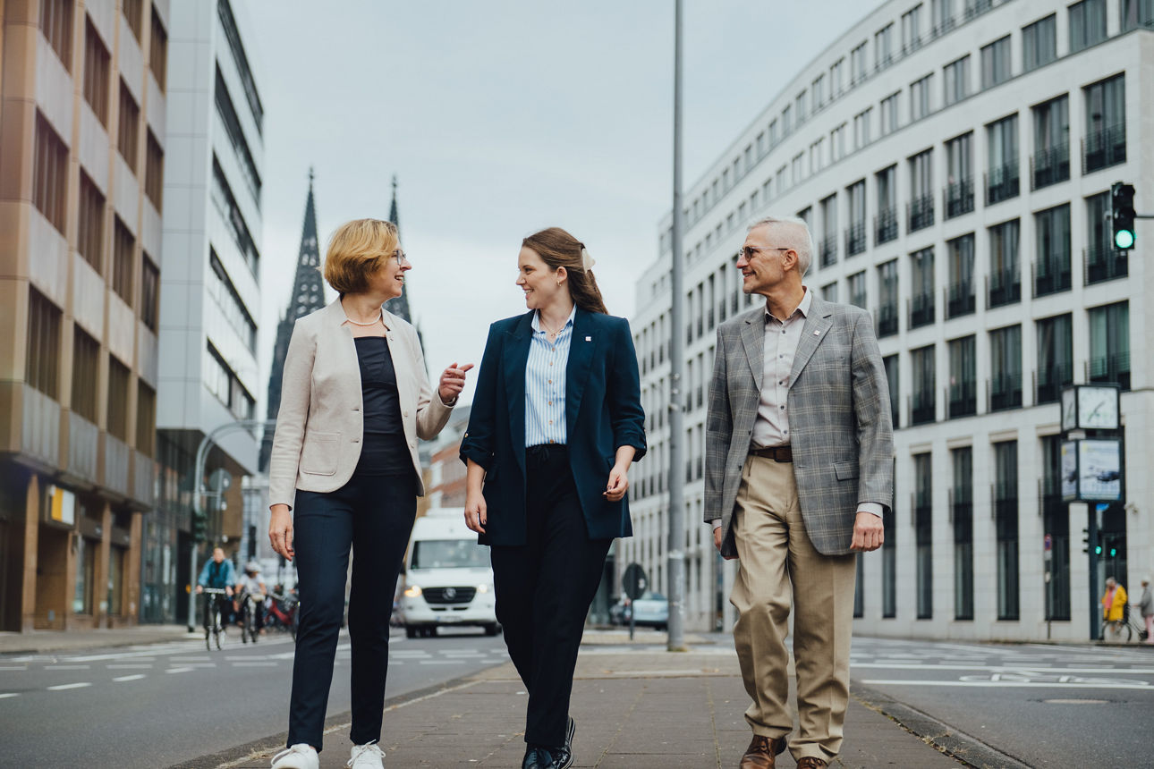 Drei Mitarbeitende laufen in der Nähe der Kölner Filiale der Pax-BKC entlang, im Hintergrund sieht man den Kölner Dom