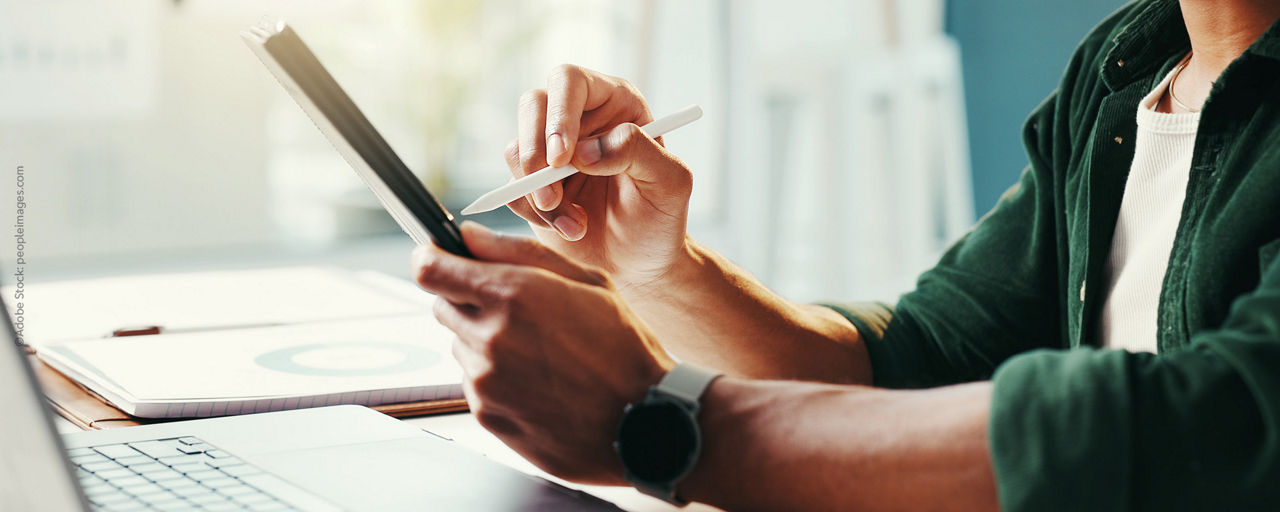 Bild zeigt zwei Hände, die eine Hand hält ein Tablet und die andere Hand ein Stift vor einem Laptop
