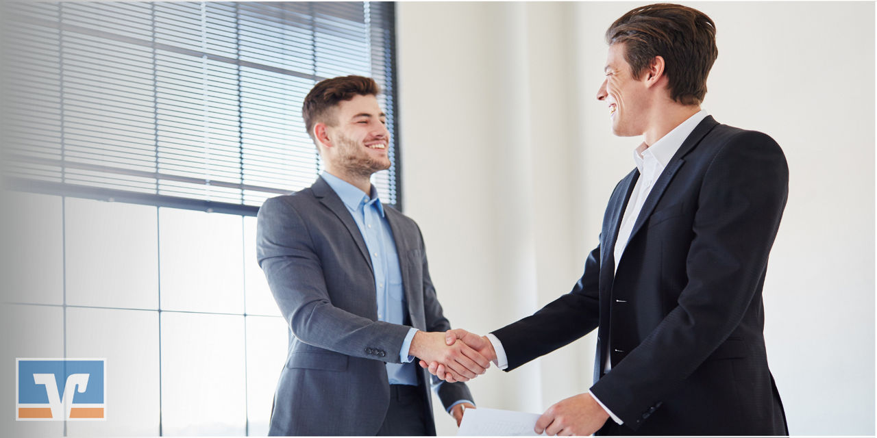 Zwei Männer in einem hellen Büro, beide in Anzügen, schütteln sich die Hände und lächeln.