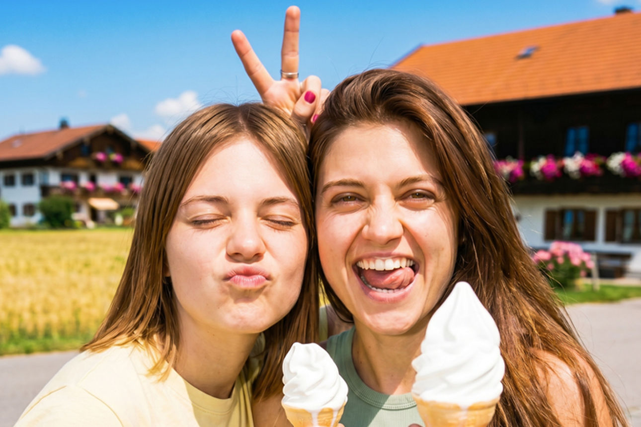 Zwei junge Frauen halten Eiswaffeln in der Hand, im Hintergrund ein bayerisches Bauernhaus.