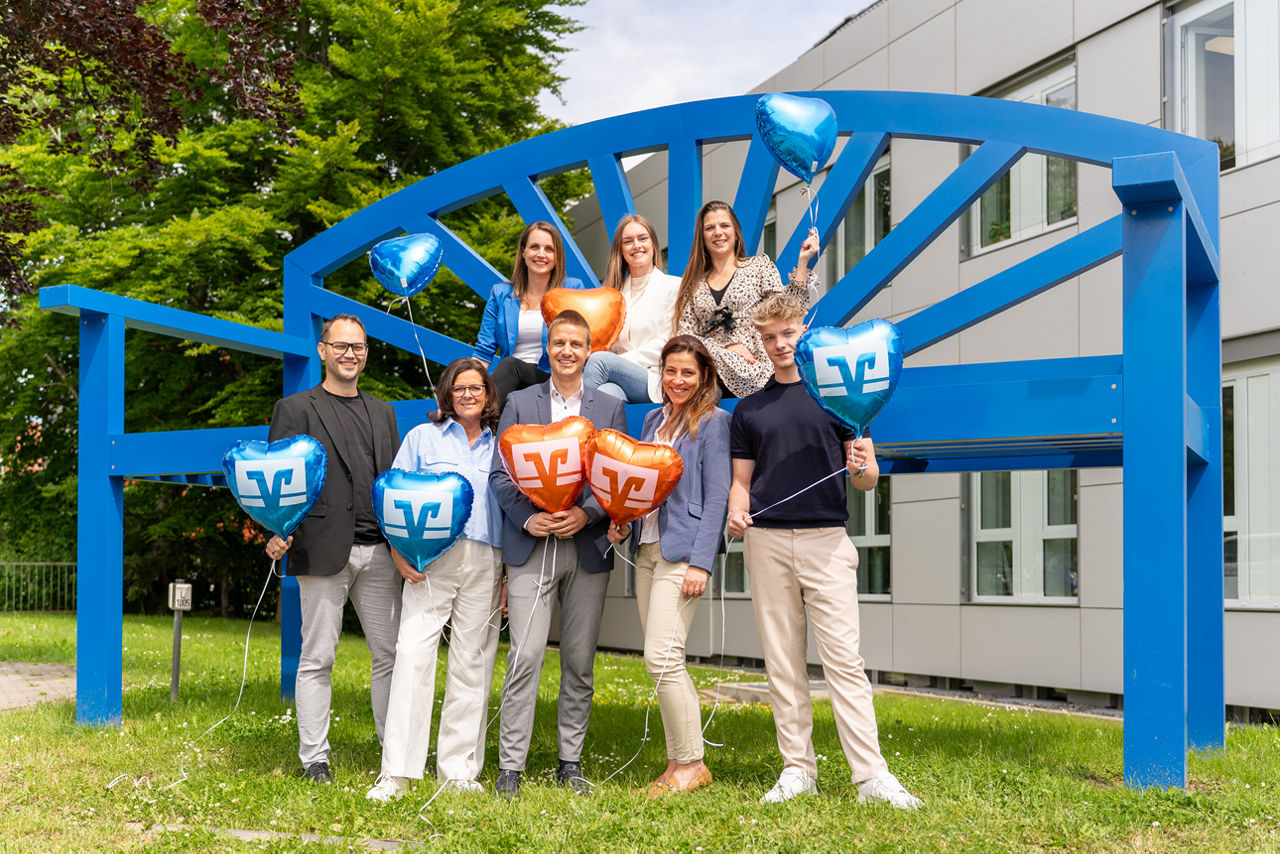 Personengruppe mit Luftballons in der Hand vor einer großen blauen Bank