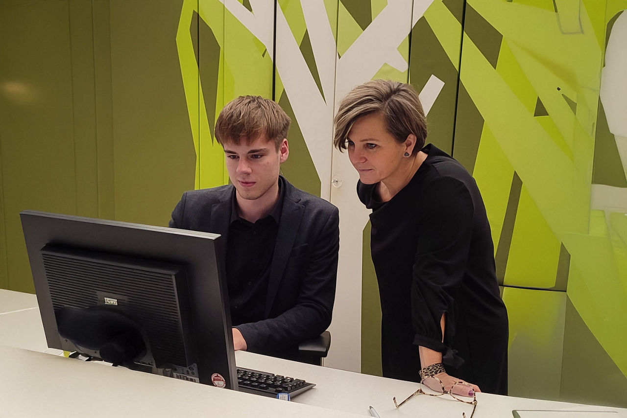 Junger Mann sitzt an einem Schreibtisch und tippt etwas. Eine Frau steht neben ihm. Beide blicken auf den Monitor.