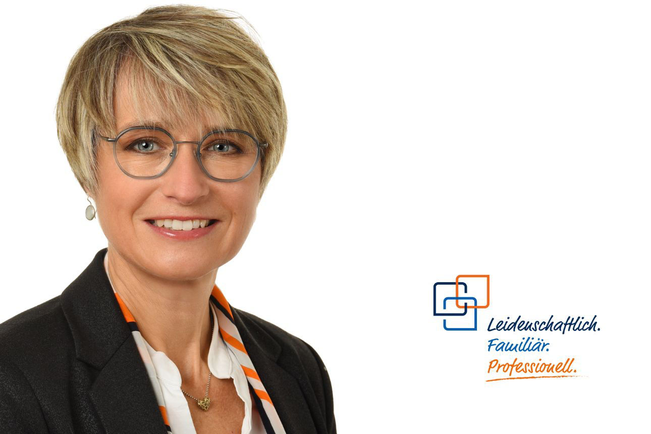 Stefanie Müller, eine Frau mit kurzen blonden Haaren, Brille und Ohrringen, lächelt freundlich und trägt eine dunkle Jacke mit einem gemusterten Schal, der orangefarbene Streifen aufweist.