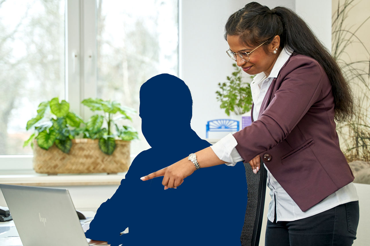 Eine Schattenfigur sitzt am Schreibtisch vor einem Laptop und eine Frau steht dahinter und zeigt auf den Bildschirm