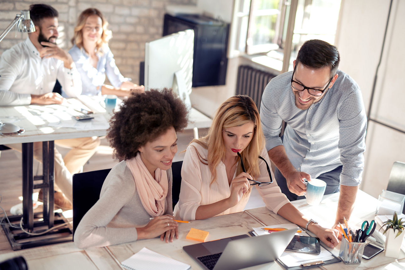 Drei Personen unterschiedlichen Geschlechts und Alters sitzen gemeinsam an einem Schreibtisch in einem hellen Büro und arbeiten konzentriert an einem Laptop und Unterlagen. Im Hintergrund zwei weitere Personen im Gespräch..