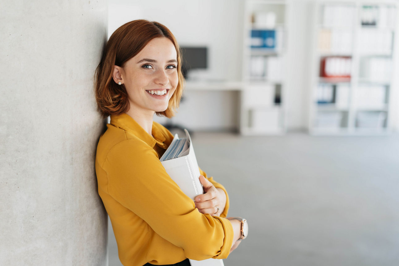 Eine junge Frau mit rotbraunen kurzen Haaren steht lächelnd an einer Wand, hält Ordner im Arm und befindet sich in einem modernen Büro.