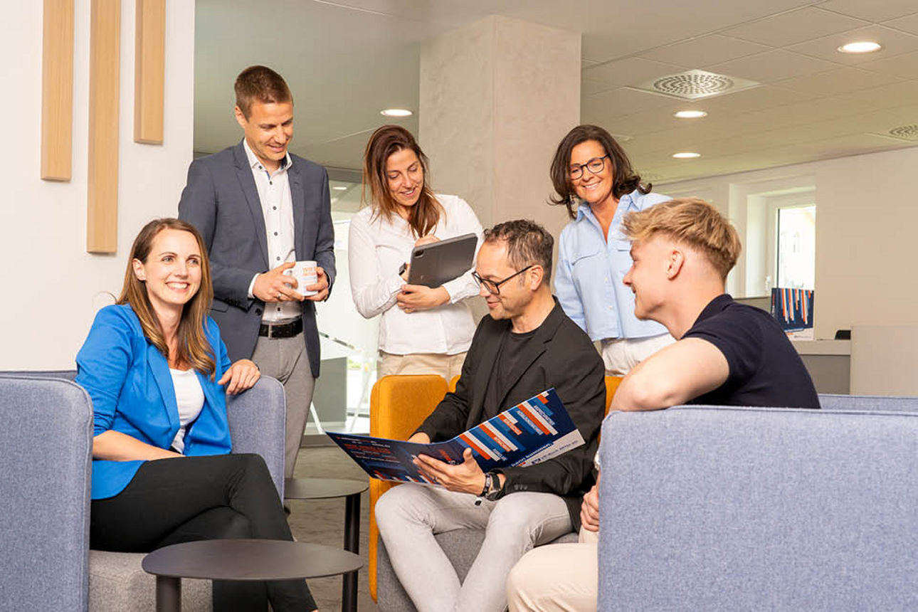 Drei Frauen und drei Männer sitzen und stehen bei einer Sitzgruppe und schauen interessiert