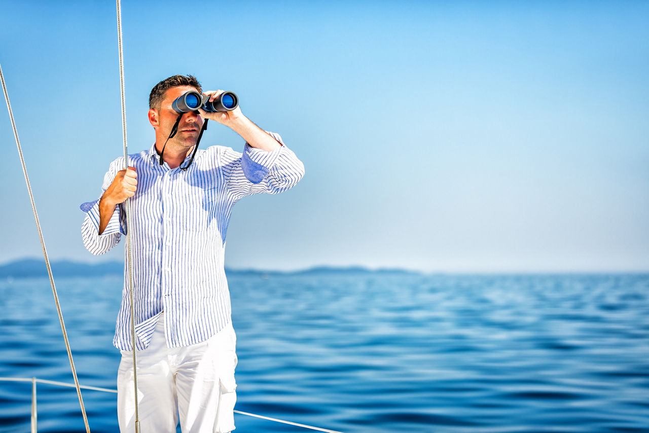 Ein Mann im Hemd steht auf einem Boot und schaut mit einem Fernglas über das Meer