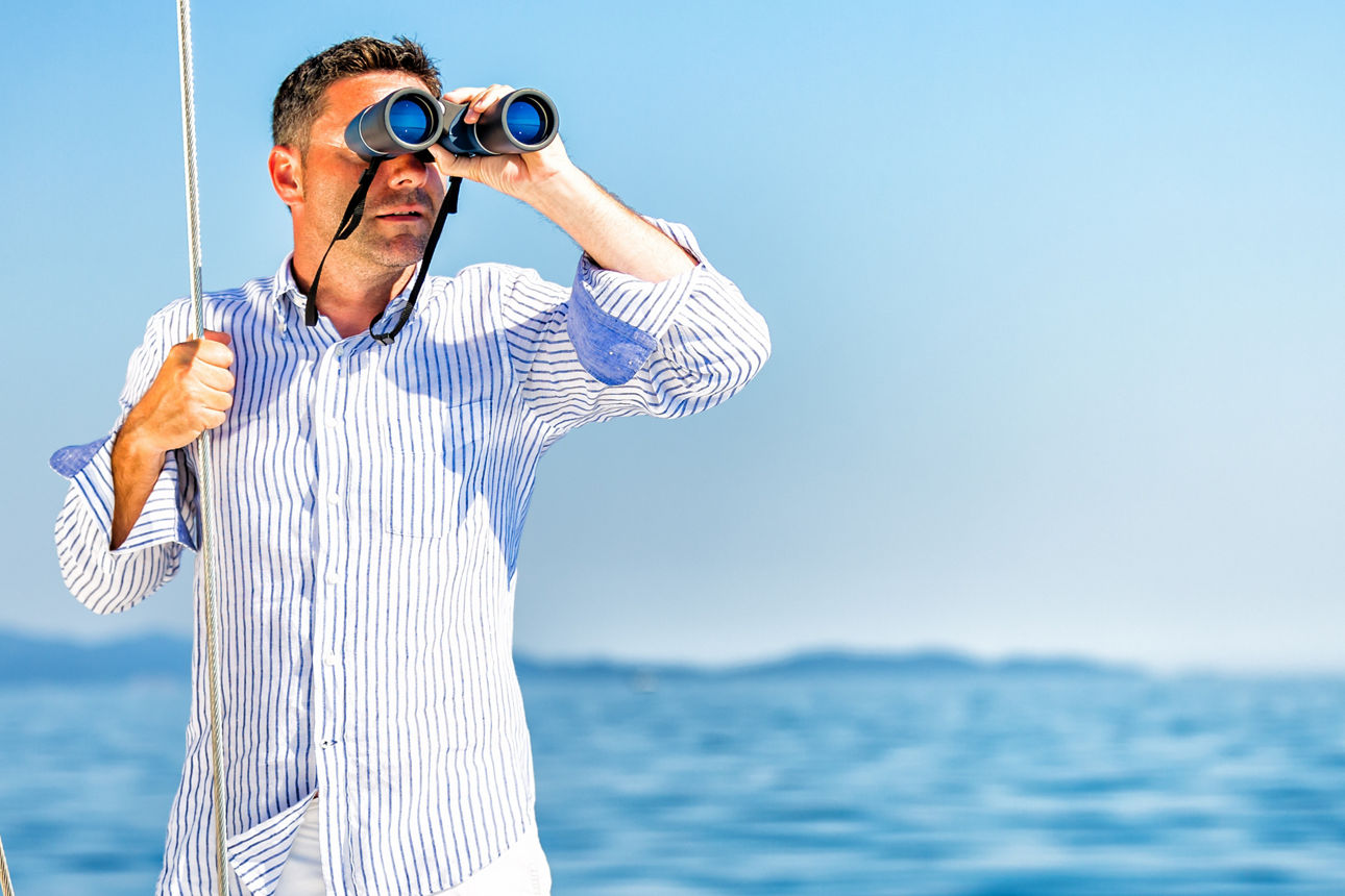 Ein Mann im Hemd steht auf einem Boot und schaut mit einem Fernglas über das Meer