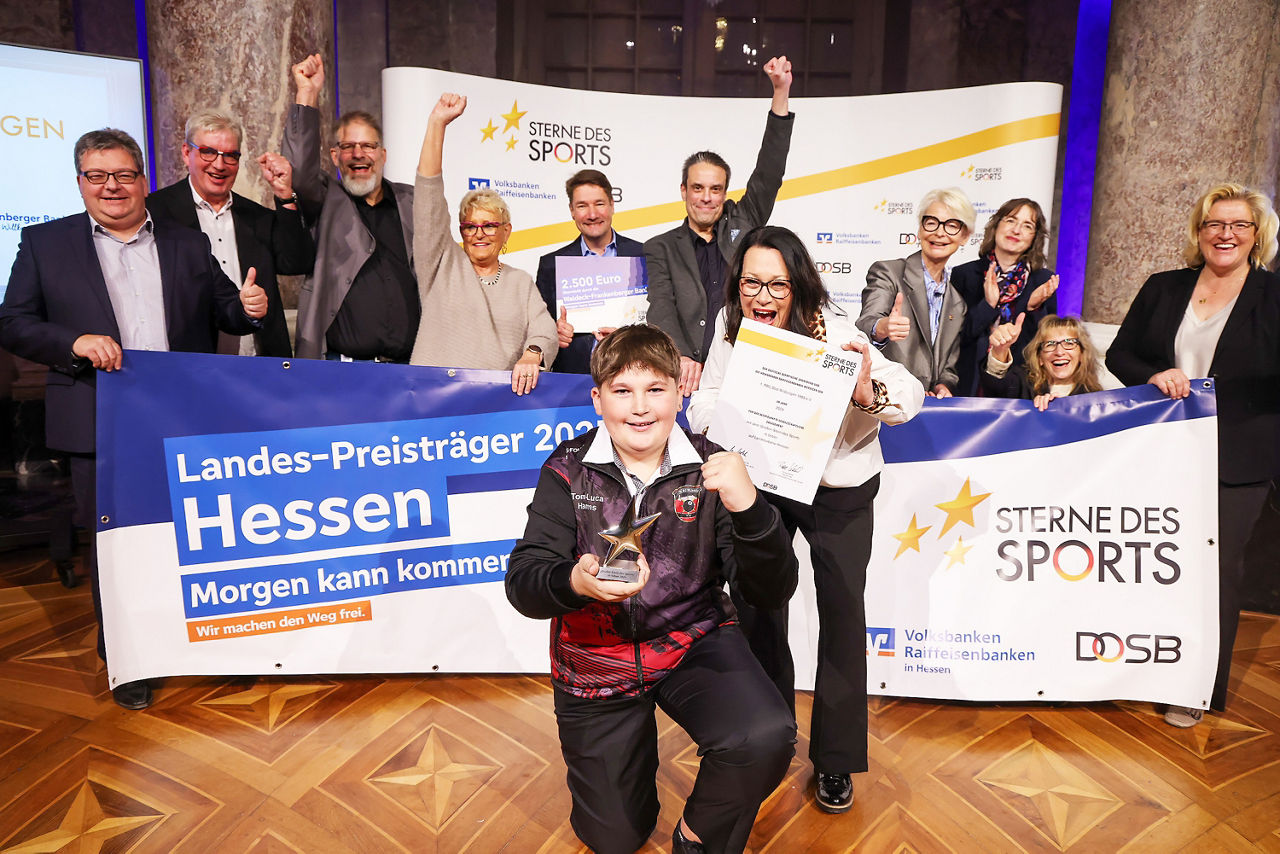 Das Bild zeigt zwölf Personen jubelnd mit einem „Sterne des Sports“ Banner. Die Gruppe setzt sich aus Mitgliedern des PBC Bad Wildungen 1989 e. V., Präsidentin des Landessportbundes Hessen, Generalbevollmächtigte der Volksbank Darmstadt Mainz eG, sowie die Sportbeauftragte des Landes Hessen