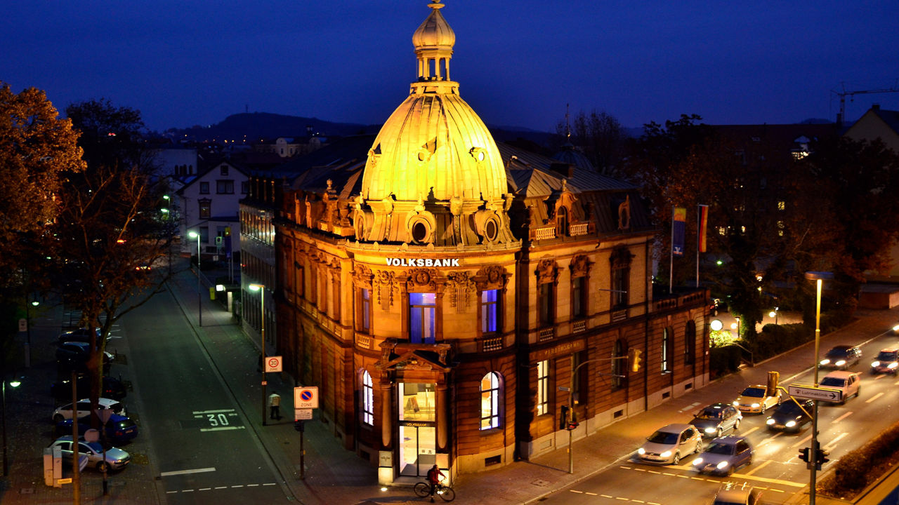 Die kunstvolle Fassade des Volksbank-Gebäudes bei Nacht, beleuchtet von warmen goldenen Lichtern.